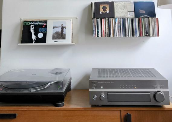 A turntable and an amplifier on top of a brown shelf.
Above them a few 7" singles and CDs
