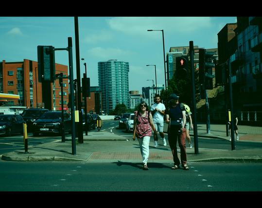 A group of pedestrians is crossing a busy urban intersection on a sunny day. At the center, a smiling woman in a red patterned top and white trousers walks beside a man in a navy shirt and red trousers, holding a bag. Behind them, other people follow, including a man in white with a beaming smile. Cars wait at the traffic lights, and modern red and glass buildings frame the background. The scene is tinted with a cool greenish-blue tone, giving it a cinematic, stylized mood. Black bars at the top and bottom add to the movie-like atmosphere.