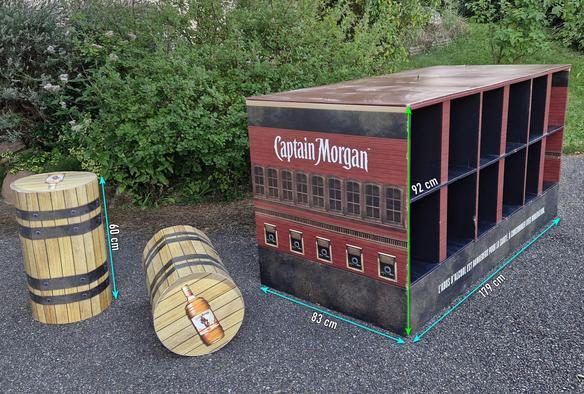 Ensemble de meubles décoratifs en carton de la marque Captain Morgan posé sur un sol en gravier, avec des arbustes verts en arrières-plan.

À gauche, deux fûts cylindriques (60 cm) en carton ressemblance "bois" avec des bandes métalliques noires, imitant les fûts de chêne traditionnels utilisés pour le stockage du rhum. 

Au centre, un grand meuble rectangulaire avec une façade imitant un bâtiment en briques rouges, orné du logo Captain Morgan. Ce meuble mesure 179 cm de long, 83 cm de large et 92 cm de haut, avec plusieurs compartiments ouverts pour ranger des bouteilles.