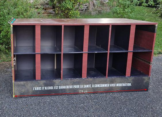 Au centre, un grand meuble rectangulaire à compartiments, probablement un distributeur de boissons. Les compartiments sont séparés par des panneaux rouges bordeau, et les intérieurs sont noirs. L'ensemble est posé sur un sol en gravier, avec des arbustes verts en arrière-plan.

Le meuble semble être en bon état, avec une finition en bois sur le dessus et des panneaux latéraux. Les dimensions sont clairement indiquées sur l'image, avec des flèches et des mesures en centimètres.

Ce meuble mesure 179 cm de long, 83 cm de large et 92 cm de haut, avec plusieurs compartiments ouverts pour ranger des bouteilles. Il est composé de dix compartiments, chacun d'une profondeur de 40 cm. 

Sur le devant du meuble, il y a une inscription en français : "L'ABUS D'ALCOOL EST DANGEREUX POUR LA SANTÉ. À CONSOMMER AVEC MODÉRATION." Cette inscription est un avertissement contre l'abus d'alcool, soulignant l'importance de la consommation modérée.