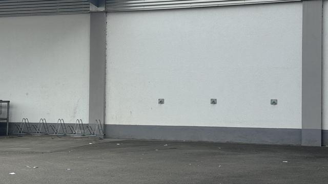 Eine leere Wand mit drei an der Wand montierten Halterungen und einer Reihe von Fahrradständern auf der linken Seite. Der Boden scheint asphaltig zu sein, mit etwas Müll verstreut.