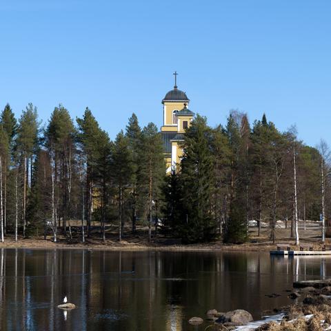 A lakeside view with a yellow church and belltower rising above a line of tall evergreen trees on the opposing shore, with a small dock in the distance. A rock with a bird sitting on it is on the lake on the right. The water is calm and reflecting the environment.