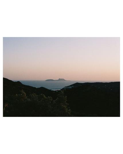 In the far distance floats an island in the soft light of sunset. In the foreground there is a strong dark contrast to the bright ocean and sky in the back. You can barely recognize leaves reflecting the sunlight. It appears as if the ocean lies in the hollow of the dark front landscape and flows off over the edge at the right side of the photo.