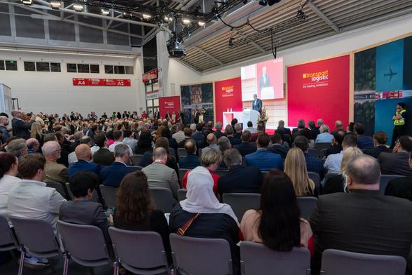 Patrick Schnieder steht vor einem Rednerpult und spricht zum Publikum. Im Hintergrund ist eine rote Rückwand zu sehen, auf der in orange und weiß folgender Text steht: "transport logistic".