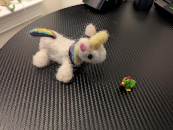 A white needle-felted unicorn with rainbow hair and a very small green chicken with red stripes.