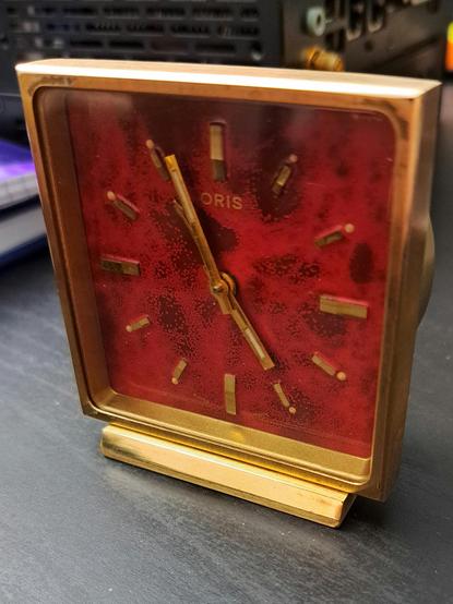 Frontal del reloj, es cuadrado con una pequeña base,  marco fino de latón, cristal de plástico y la esfera es roja brillante con manchas hechas adrede.

Tiene 3 agujas, minutos, horas y la aguja del despertador.