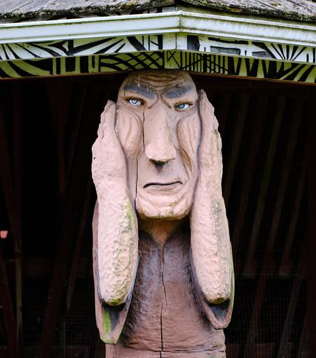 A carving of a troll, actually one of the posts holding up to roof of a barn on the Troll Haven estate on the Olympic Peninsula.  Its hands are covering its ears and has a furroughed brow.  The gutter and shingles on the roof can just be seen at the top of the photo.  Other carvings throughout of dragons, other trolls etc.