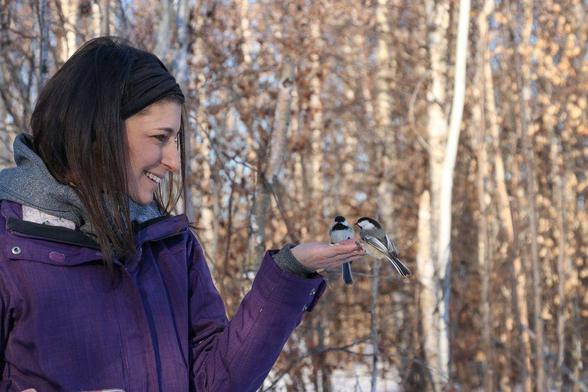 Photo of myself in early winter with hand slightly outstretched with some wild birdseed in my palm. Two chickadees are perched on my finger tips, and I am smiling in delight. A mix of birch and aspen with brown leaves are in soft focus in the background.