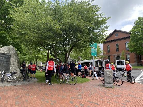 Day 1. A gathering of cyclists at a park in Concord, MA. There are red and grey bricks that perimeter a grassy and tree-inhabited park, and there is a stone-encased drinking water fountain. There are about 20 cyclists, most wearing a red-and-white jersey that says Bostréal 2025 on it, and they are socializing and getting snacks out of a white van that is parked adjacent to the park.