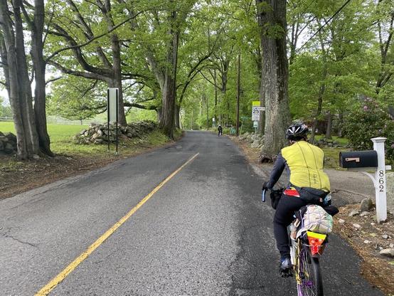 Day 1. A cyclist wearing a yellow and grey jacket, black pants, and riding a yellow bike with purple rims is riding on a rural road that a single yellow line down the middle for a little distance before a town line when the yellow line ends. In the distance are two other cyclists. This is at the edge of Concord, MA and Carlisle, MA on Strawberry Hill Rd.