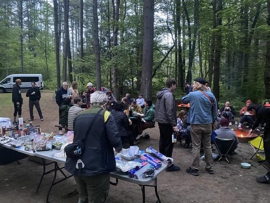 Day 1. An outdoor campsite in Otter River State Forest that has been set up as a group dinner location. There are over 20 people sitting and standing around the food tables, campfire, camp grill, and in areas to socialize and chat. The food table has an assortment of prepared, uncooked food and aluminum foil which acted as both cooking vessel for the grill and bowl/plate.