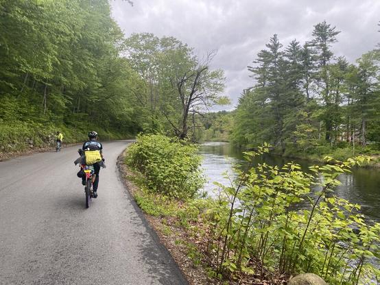 Day 2. A cyclist wearing a black HUP kit jersey, yellow and grey jacket, black pants, and riding a yellow bike with purple rims is riding on a rural road and waving to a cyclist wearing high-visibility jacket and light blue bike going in the opposite direction. There are forest trees to the left of the road and the Ashuelot River to the right.