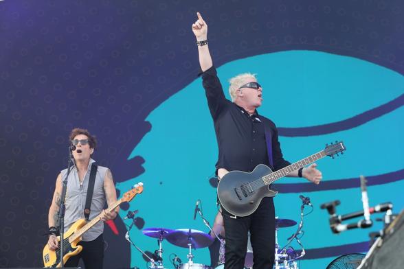 The Offspring perform on stage in 2022; from left: bassist Todd Morse, and guitarist and singer Dexter Holland who is brandishing his guitar with his strumming hand pointing to the sky.
