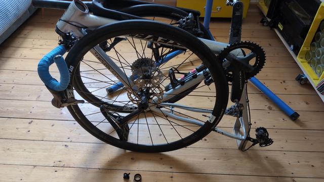 Ein zerlegtes Fahrrad. Im Hinterbau ist ein kleiner Ständer montiert, sodass das Rad darauf und auf dem Sattel (quasi auf dem Rücken) liegt. Die Räder sind herausgenommen und lehnen von beiden Seiten am Rahmen. Die Gabel liegt oben auf dem Unterrohr und der Lenker baumelt an der Seite.