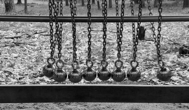 Text von ChatGPT:

Das Bild zeigt eine Schwarz-Weiß-Aufnahme von einer Reihe metallischer Kugeln, die an robusten Ketten aufgehängt sind. Die Kugeln sind in einer Linie angeordnet und scheinen Teil eines großen Pendelsystems zu sein, das an Newtons Wiege erinnert. Im Hintergrund ist ein herbstlicher Waldboden mit fallenden Blättern und Bäumen zu sehen. Eine Kugel auf der rechten Seite ist in Bewegung und schwingt nach außen, während die anderen Kugeln ruhig hängen. Die Ketten sind an einer stabilen Metallstange befestigt, die quer über das Bild verläuft.