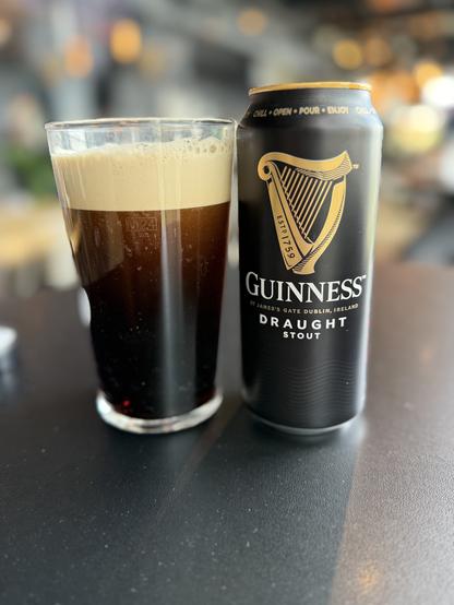 A pint glass with a proper nitro pour settling is next to a black pint can with the gold Guinness harp logo