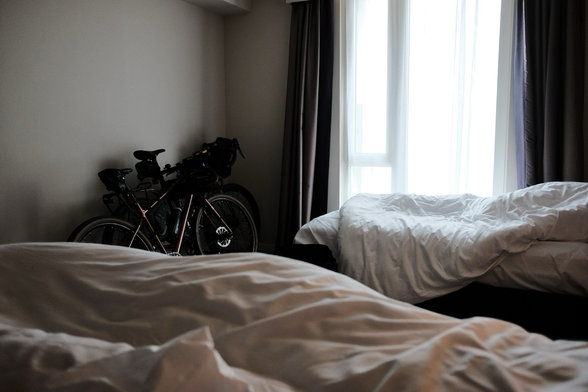 Two bikes lean against the wall of a hotel room, unmade beds in the foreground.