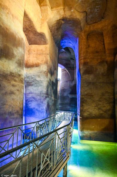 Blick in einen historischen Wasserspeicher in Süditalien, das für Besucher geöffnet ist. Ein langer Gang führt durch das hohe unterirdische Gewölbe, das von Scheinwerfern bunt beleuchtet wird. /
A view of a historic water reservoir in southern Italy, open to visitors. A long walkway leads through the high underground vault, which is colorfully illuminated by spotlights.