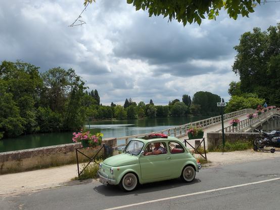 Photo centrée sur une petite voiture Fiat 500 couleur vert pastel, en bord de Seine, derrière elle se profile un pont pour piétons avec une balustrade en bois blanc et des jardinières ornées de géraniums roses. Le ciel est gris, couvert, mais le soleil est quand même là