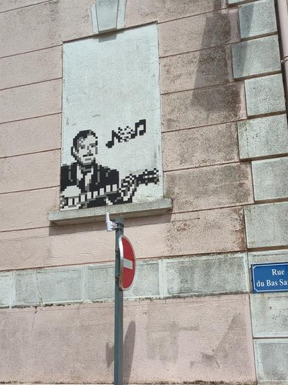 Photo d'un mur beige, dans une fausse fenêtre se trouve une mosaïque représentant un homme moustachu en noir et blanc en train de jouer de la guitare à la fenêtre. Au dessus de lui, quelques notes de musique noires dissimulent un personnage de space invader blanc