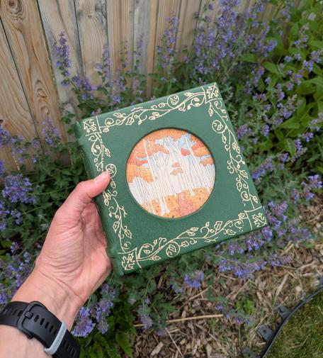 A square, green leather journal that has a round painting of a forest in the center of the cover. Surrounding the painting is a drawing of gold vines.