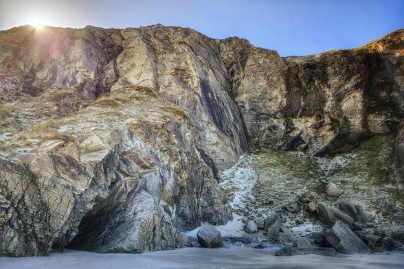 A stunning view of some towering cliffs in County Donegal, Ireland. The cliffs rise dramatically from the beach sand on the Atlantic coast, showcasing their immense height and rugged, layered rock formations. A dark cave entrance can be seen at the bottom left of the wall of rock. The sky is a blue hue with the bright sun just about to disappear behind the cliffs on the left.