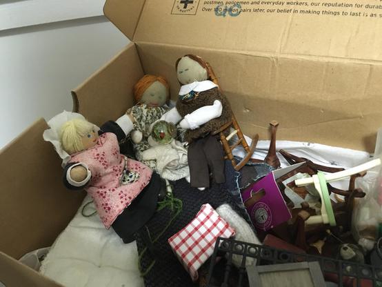 Three cloth dolls, a man, a woman and a child doll in a cardboard box with a wooden doll wearing a white cap and a wrap-around pink apron. Furniture, cushions and other bits of their possessions are scattered about them.