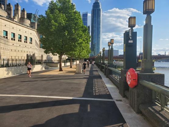 Footpath along the Thames behind a large cream coloured building. There are several runners on the wide path.