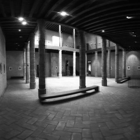 Fotografía en blanco y negro del interior del primer piso del palacio del Condestable. En la calle Mayor del casco antiguo de Pamplona. La fotografía es oscura aunque se ven los diferentes puntos de luz del techo y de la entrada central de luz natural del tejado. Se ve en primer lugar un banco muy simple. Toda la sala está vacía salvo que hay fotografías pequeñas en las paredes y supongo que hice la foto con un angular ya que se ve un poco deformada.