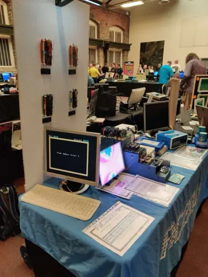The RC2014 stall in an exhibition hall. There's a blue tablecloth with the RC2014 logo on, and a stepped platform in the middle with different RC2014 models on it. The nearest monitor is showing the TMSNake game from Wintermute. Behind that is a bright LED representation of a Mandlebrot fractal. Towards the far end of the table is another monitor and an RC2014 in a Blue Box next to the Etch-a-Sketchy display.