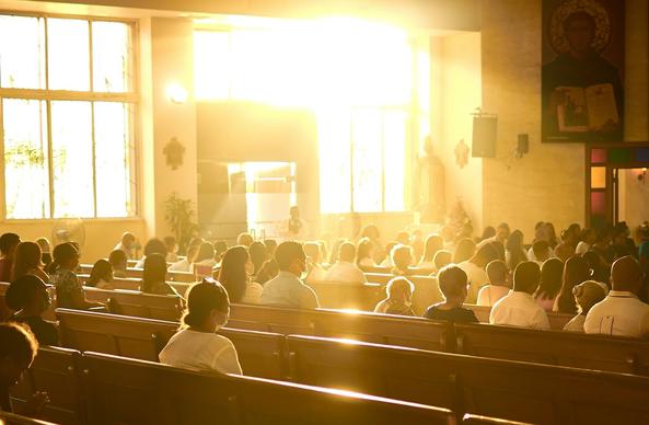 Bright yellow sunlight streams through the windows of a house of worship, illuminating the people sitting in their pews.