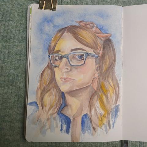 An open sketchbook with mostly out of view a blank right page. On the left page is a portrait painted in watercolour paint. The woman is white, her face is turned towards the viewer's left, but she looks at the viewer. The woman has a salmon pink bow in her hair and two hair clips in the same colour. Her hair is past her shoulders and wavy painted in brown and yellow splotchy colours. She's wearing a blue jacket and the background is painted in a paler hue of the same colour.
There's a golden paper clamp on the top left of the left page.