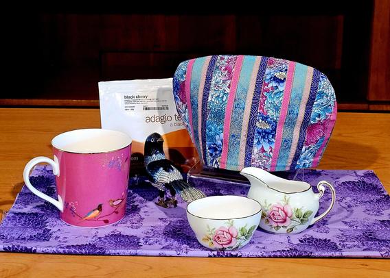 Tea tableau in deep purple and bright pink with an Asian inspired tea cozy, chrysanthemum patterned tea towel, delicate vintage rose-print cream and sugar, a Portmeirion Sarah Miller mug with tropical birds, and a cloisonne bird statue. The tea is Black Cherry black loose-leaf from Adagio.