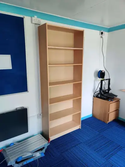 The back wall of RC2014 where a wooden bookshelf sits between a blue notice board and a small set of drawers with a 3D printer on them.