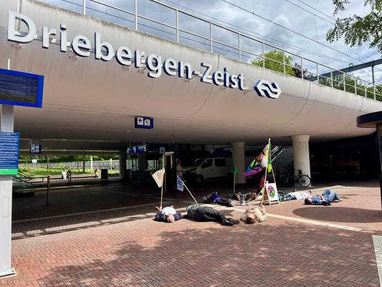 Station Driebergen-Zeist. Naast het station ligt een groep rebellen op de grond, alsof ze dood zijn. Sommige rebellen houden XR vlaggen vast.