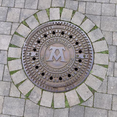 Runder, mit einem Ring aus Steinen umfasster Schachtdeckel aus Metall. Im Zentrum befindet sich das Symbol der Formel "Mons, Pons, Fons" (Berg, Brücke, Quelle), die die Entwicklung der Stadt Lüneburg beschreibt. Um das Symbol herum stehen die beiden Worte "Lüneburg" und "Stadtentwässerung".
