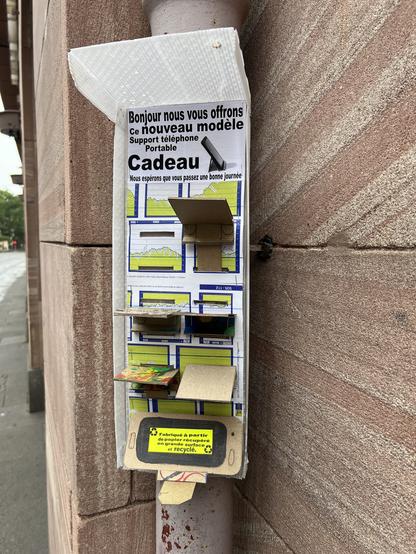 Accroché à une gouttière verticale dans une rue du centre ville de Strasbourg, un petit présentoir en carton sur lequel est écrit «&nbsp;Bonjour nous vous offrons ce nouveau modèle Support téléphone Portable. Cadeau. Nous espérons que vous passez une bonne journée&nbsp;». Accompagné d’une photo d’un téléphone portable posé sur un petit support. En dessous du message une série d’encoches dans lesquelles sont enfichés des morceaux de carton pliés que tout le monde peut prendre et qui semblent être les supports en question. Ils sont tous décorés différemment.
Tout en bas un autre message indique «&nbsp;Fabriqué à partir de papier récupéré en grande surface et recyclé&nbsp;»
Le tout semble être protégé sur les côtés par une matière plastique de sorte à éviter d’être trempé par la pluie.