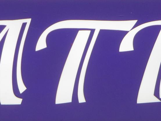 Белая буква Т на фиолетовом фоне, часть слова-вывески «Ателье». // White letter T on a violet background, part of the word sign “Atelier”.