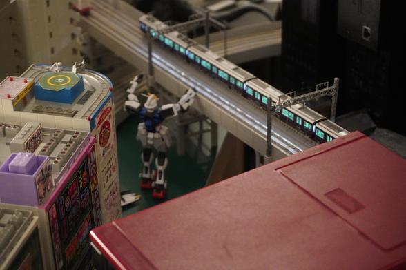 Areal shot of a city, a gundam watching over the trains speeding trough an overpass