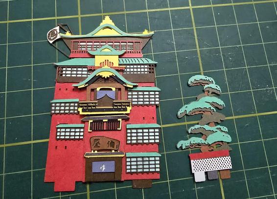 A tiny paper craft model in progress of the very magical bath house in the movie "Spirited Away", resting on a rubber cutting mat.