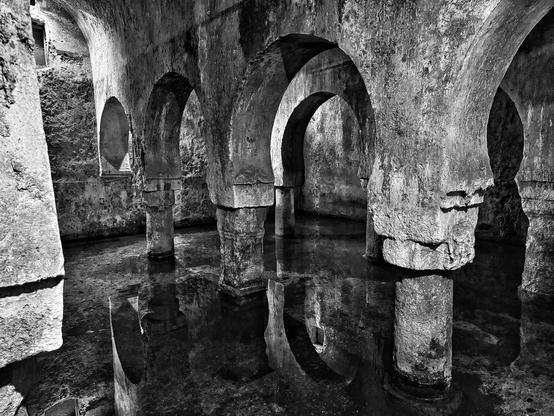 Fotografía en blanco y negro de una cámara subterránea con múltiples columnas de piedra que sostienen arcos. El agua cubre el suelo, reflejando las columnas y los arcos de manera simétrica. Las paredes de piedra muestran signos de desgaste y antigüedad. La escena evoca una atmósfera misteriosa y antigua.
