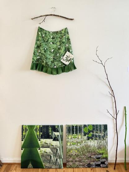 On the wall is hanging a skirt made of textile with forest floor pattern. The skirt is hanging on a hanger made by a stick found in the forest.
On the floor below are 2 pictures made of digital print on canvas and frame depicting kaleidoscopic images from a bog in Jægersborg Hegn in Copenhagen. 
Next to the pictures are 2 sticks, a green and a natural, standing, leaning against the wall.