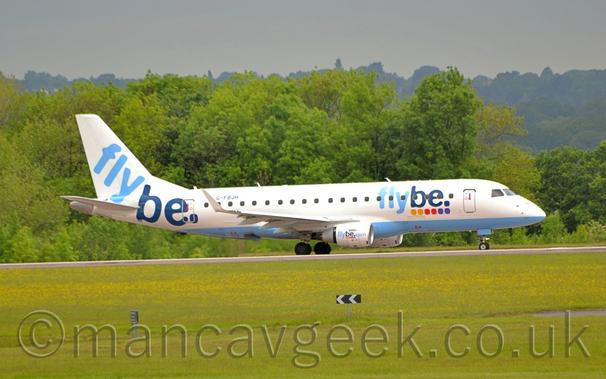 Side view of a white, twin engined jet airliner with the engines mounted on the sides of the rear fuselage, flying from left to right at a very low altitude, maybe a metre above as runway, with the undercarriage lowered, flaps extended from the rear of the wings, and the nose fairly flat, suggesting it is about to land.
Green grass fills the foreground, lining the side of the runway, with more grass in the background, leading up to trees in the background.
The rather dilapidated white fuselage of a 3 engined jet airliner is visible in front of the trees, facing to the left.
Grey sky fills the rest of the frame.