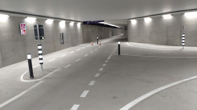 Blick auf eine T-Keuzung mit ungewohnt schmalen Fahrbahnen und winzigen Verkehrsinselnin einen modernen, hell erleuchteten Tunnel aus Beton. An der Wand eines der typischen, weinroten Schweizerischen Velowegschilder.