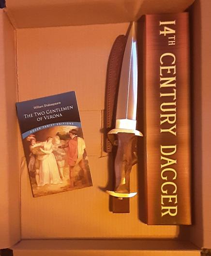 Inside of an opened cardboard box is a cooy of Shakespeare's "Two Gentlemen of Verona" and a 15 inch medieval dagger with a wooden handle, out of its shipping box. There is also a leather sheath.