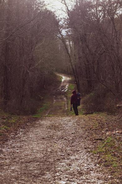 Image of a man hiking along a muddy trail in the woods. The trees are mostly bare with spots of grass appearing on the ground in early Spring.