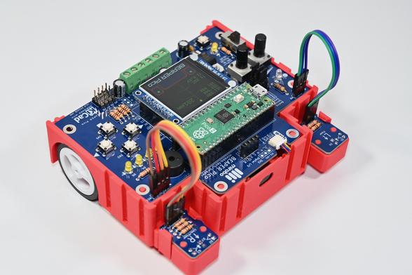 Front-right view of a red 3D-printed BEAPER Bot robot controlled by a Raspberry Pi Pico 2 microcontroller mounted in a blue BEAPER Pico printed circuit board. The LCD display mounted behind the Raspberry Pi Pico is displaying a radar-like display showing the distance to an object sensed by a time-of-flight (ToF) module mounted in the oval window at the front, and the reflectivity of the floor sensed by the left and right optical floor sensor modules attached by rainbow-coloured extension cables.