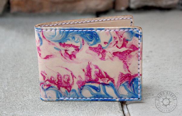 The front of a handmade leather wallet. It is natural leather with blue and pink marbling resembling a trans flag. It has white stitching