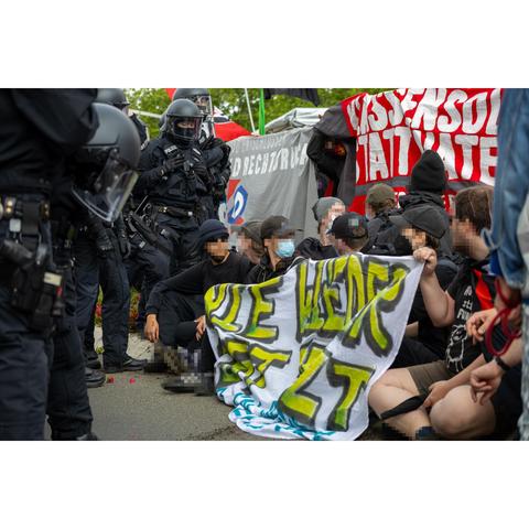 Sitzblockade. Dahinter sind mehrere banner. Gegenüber stehen mehrere behelmte Cops.