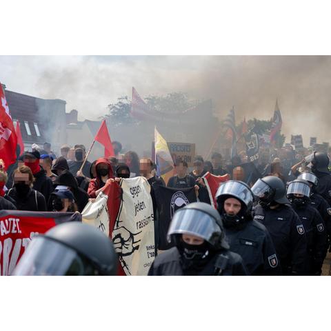 Demontrationszug. Mehrere Banner sind zu sehen. In der Demo wurde Pyro gezündet. Rauch steigt aus der Demo auf. Neben der Demo laufen behelmte Cops